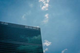 A modern office building with reflective glass windows is positioned against a clear blue sky dotted with a few white clouds.