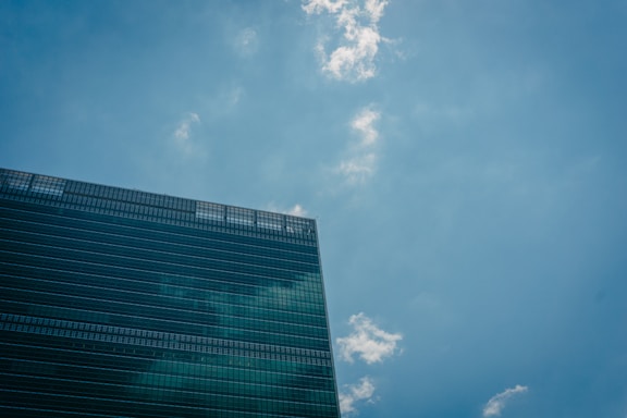 A modern office building with reflective glass windows is positioned against a clear blue sky dotted with a few white clouds.