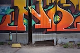 A colorful graffiti mural on a wooden door features bold geometric shapes and vibrant colors, with a yellow broom leaning against the wall near a spray paint can. The door is slightly open, revealing a dark interior.
