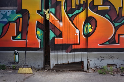 A colorful graffiti mural on a wooden door features bold geometric shapes and vibrant colors, with a yellow broom leaning against the wall near a spray paint can. The door is slightly open, revealing a dark interior.
