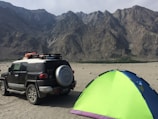 A rugged outdoor scene with a sport utility vehicle parked on a sandy terrain next to a brightly colored tent. The background features towering, rocky mountains under a clear sky.