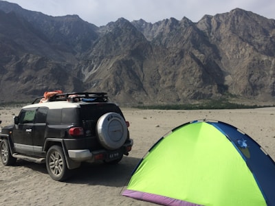 A rugged outdoor scene with a sport utility vehicle parked on a sandy terrain next to a brightly colored tent. The background features towering, rocky mountains under a clear sky.