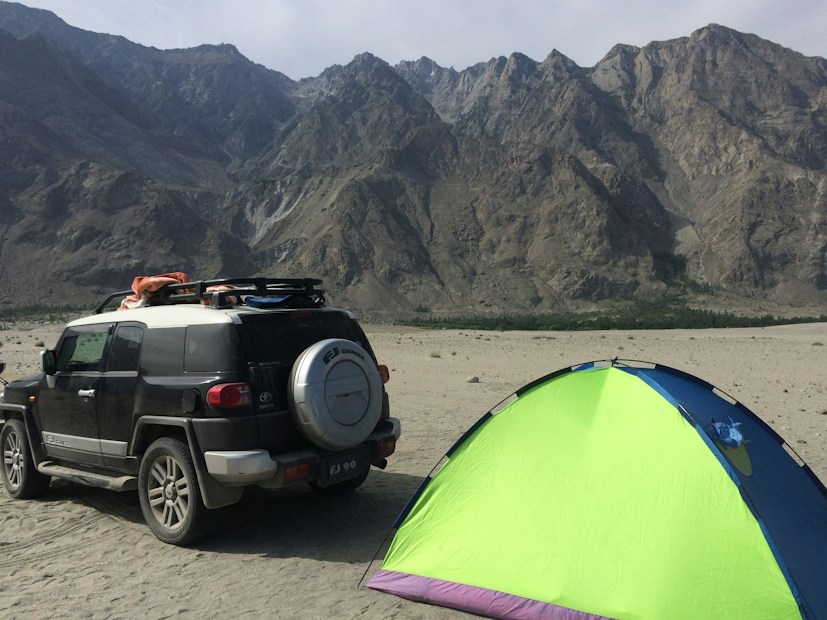 A rugged outdoor scene with a sport utility vehicle parked on a sandy terrain next to a brightly colored tent. The background features towering, rocky mountains under a clear sky.
