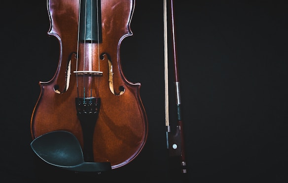 A close-up of a violin next to a bow on a dark background. The violin's body displays a rich, polished wood texture, with visible strings and fine detailing in the design.