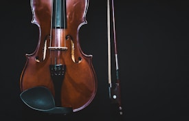 A close-up of a violin next to a bow on a dark background. The violin's body displays a rich, polished wood texture, with visible strings and fine detailing in the design.