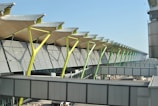 A modern architectural structure with a series of slanted, colorful support beams under a roof with a wavy design. The building features large windows and an exterior with horizontal metal panels. A walkway or bridge connects parts of the structure, with urban elements visible in the surrounding area.