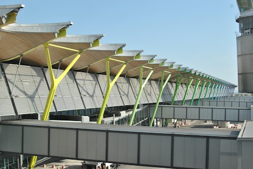 A modern architectural structure with a series of slanted, colorful support beams under a roof with a wavy design. The building features large windows and an exterior with horizontal metal panels. A walkway or bridge connects parts of the structure, with urban elements visible in the surrounding area.