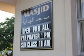 A sign on a building wall displays information in both Arabic and English. It announces 'Masjid' along with the message 'Open for All, Fri Prayer 1 PM, Sun Class 10 AM'. The lettering is white against a black background. Part of the building and window are visible, along with greenery outside.