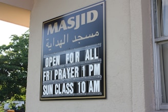 A sign on a building wall displays information in both Arabic and English. It announces 'Masjid' along with the message 'Open for All, Fri Prayer 1 PM, Sun Class 10 AM'. The lettering is white against a black background. Part of the building and window are visible, along with greenery outside.