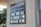 A sign on a building wall displays information in both Arabic and English. It announces 'Masjid' along with the message 'Open for All, Fri Prayer 1 PM, Sun Class 10 AM'. The lettering is white against a black background. Part of the building and window are visible, along with greenery outside.