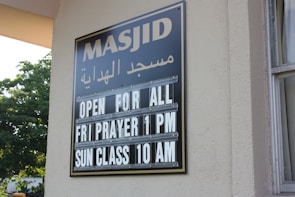 A sign on a building wall displays information in both Arabic and English. It announces 'Masjid' along with the message 'Open for All, Fri Prayer 1 PM, Sun Class 10 AM'. The lettering is white against a black background. Part of the building and window are visible, along with greenery outside.