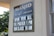 A sign on a building wall displays information in both Arabic and English. It announces 'Masjid' along with the message 'Open for All, Fri Prayer 1 PM, Sun Class 10 AM'. The lettering is white against a black background. Part of the building and window are visible, along with greenery outside.