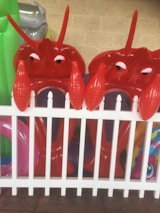 Two inflatable red lobster toys are positioned behind a white picket fence. Bright and colorful pool inflatables are visible in the background, adding to a playful atmosphere. The setting appears to be outdoors, possibly in a yard or near a pool.