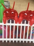 Two inflatable red lobster toys are positioned behind a white picket fence. Bright and colorful pool inflatables are visible in the background, adding to a playful atmosphere. The setting appears to be outdoors, possibly in a yard or near a pool.