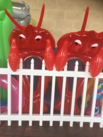 Two inflatable red lobster toys are positioned behind a white picket fence. Bright and colorful pool inflatables are visible in the background, adding to a playful atmosphere. The setting appears to be outdoors, possibly in a yard or near a pool.