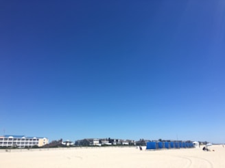 A vast expanse of sandy beach stretches across the foreground with clear blue skies overhead. Rows of blue beach cabanas are lined up on the right side of the beach. Buildings with white facades and blue roofs are visible in the distance, indicating a coastal setting.