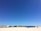 A vast expanse of sandy beach stretches across the foreground with clear blue skies overhead. Rows of blue beach cabanas are lined up on the right side of the beach. Buildings with white facades and blue roofs are visible in the distance, indicating a coastal setting.
