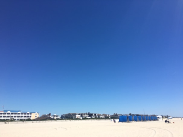 A vast expanse of sandy beach stretches across the foreground with clear blue skies overhead. Rows of blue beach cabanas are lined up on the right side of the beach. Buildings with white facades and blue roofs are visible in the distance, indicating a coastal setting.