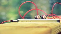 A smartphone rests on a wooden surface with a pair of red and silver earphones placed on top. The earphones have red cables that loop around the device. The background is blurred, creating a bokeh effect with soft green tones.