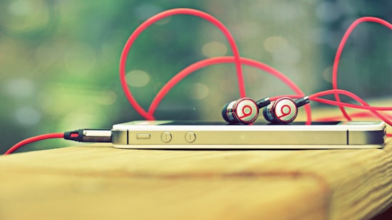 A smartphone rests on a wooden surface with a pair of red and silver earphones placed on top. The earphones have red cables that loop around the device. The background is blurred, creating a bokeh effect with soft green tones.