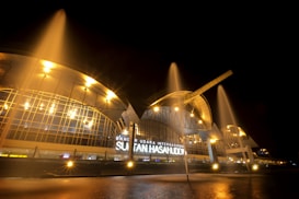 An illuminated modern architectural structure with glass and metal elements is visible at night. Bright artificial lights create a dramatic effect, and water fountains are prominently displayed, arching towards the sky. The name 'Sultan Hasanuddin International Airport' is visible on the building.