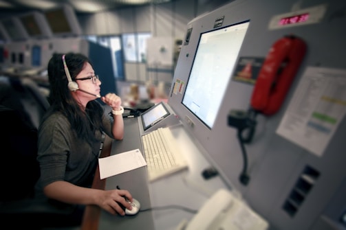 A person wearing glasses and a headset is seated at a large control panel, focusing on a computer screen. The panel includes various equipment such as a red phone and several control buttons. Papers and a keyboard are arranged in front of them, and the environment appears to be a control room.