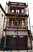 An old, three-story building is reinforced with red scaffolding. The structure has weathered walls, peeling paint, and closed metal shutters on the ground floor. The windows on the upper floors are framed in blue and red, surrounded by faded yellow walls. Overhead wires and fencing enclose the building.