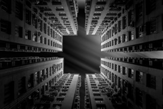 A black and white photograph taken from the ground, looking up at the symmetrical sides of tall residential buildings. The buildings have numerous windows and balconies, creating geometric patterns. The view opens to the sky, revealing a cross-shaped open space with dramatic lighting effects.