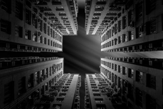 A black and white photograph taken from the ground, looking up at the symmetrical sides of tall residential buildings. The buildings have numerous windows and balconies, creating geometric patterns. The view opens to the sky, revealing a cross-shaped open space with dramatic lighting effects.