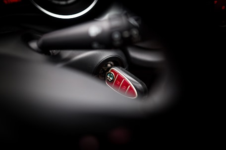 A close-up view of a Mini car key fob inserted in the ignition, with blurred surrounding elements of the car's interior. The key fob has a shiny, red surface with several buttons.