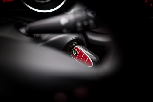 A close-up view of a Mini car key fob inserted in the ignition, with blurred surrounding elements of the car's interior. The key fob has a shiny, red surface with several buttons.