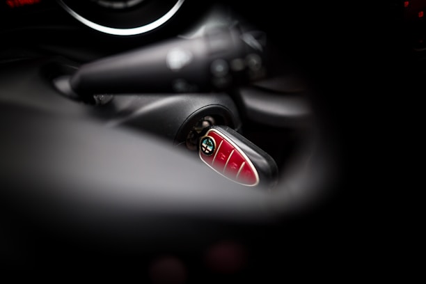 A close-up view of a Mini car key fob inserted in the ignition, with blurred surrounding elements of the car's interior. The key fob has a shiny, red surface with several buttons.