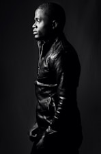 A person is captured in profile view, wearing a leather jacket. The monochrome image highlights the textures of the jacket and the subject's hair. The lighting creates dramatic shadows, accentuating the contours of the face and the clothing, contributing to a strong contrast against the dark background.