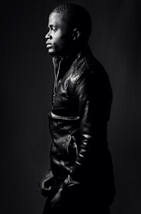 A person is captured in profile view, wearing a leather jacket. The monochrome image highlights the textures of the jacket and the subject's hair. The lighting creates dramatic shadows, accentuating the contours of the face and the clothing, contributing to a strong contrast against the dark background.