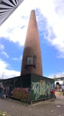 A tall brick chimney stands in an urban setting, surrounded by a structure covered with graffiti art. The sky is bright with scattered clouds. There are two people near the base of the chimney, one of whom is visible and appears to be engaged in painting or graffiti. Metal poles, a ladder, and street lights are also visible, along with a building and some trees in the background.