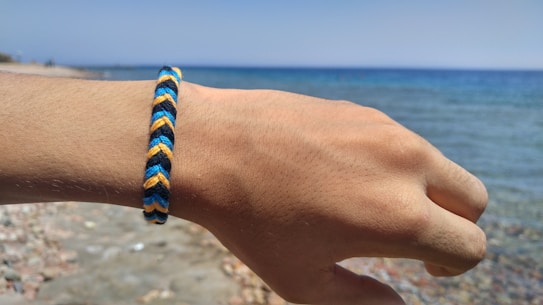 A hand wearing a woven friendship bracelet with blue, yellow, and black colors. The background features a beach scene with a pebbly shore and a clear blue sea under a bright sky.