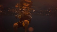 A scuba diver is submerged in dark water, partially illuminated by faint golden light reflecting from the surface. The visibility is low, creating a moody and mysterious atmosphere. The diver's legs and part of the torso are visible, and they appear to be wearing a wet suit with yellow accents.