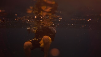 A scuba diver is submerged in dark water, partially illuminated by faint golden light reflecting from the surface. The visibility is low, creating a moody and mysterious atmosphere. The diver's legs and part of the torso are visible, and they appear to be wearing a wet suit with yellow accents.
