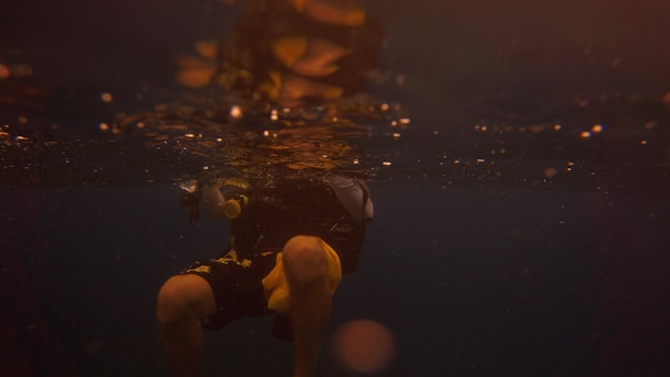 A scuba diver is submerged in dark water, partially illuminated by faint golden light reflecting from the surface. The visibility is low, creating a moody and mysterious atmosphere. The diver's legs and part of the torso are visible, and they appear to be wearing a wet suit with yellow accents.