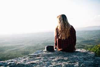 woman wearing red and black gingham shirt sitting on cliff with hat by its side