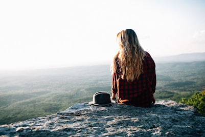 woman wearing red and black gingham shirt sitting on cliff with hat by its side