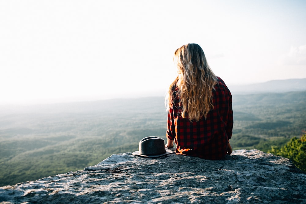 woman wearing red and black gingham shirt sitting on cliff with hat by its side