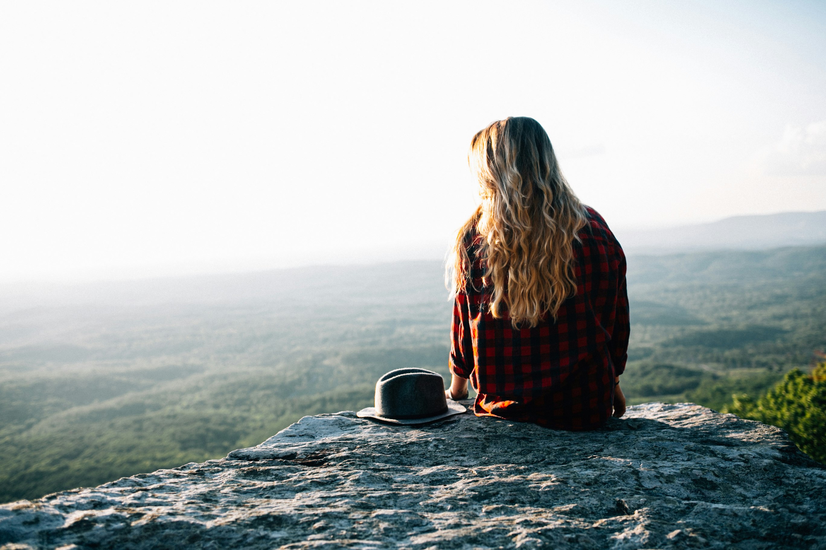 woman wearing red and black gingham shirt sitting on cliff with hat by its side