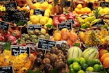 An assortment of colorful and exotic fruits displayed in baskets at a market. Each type of fruit is labeled with a small chalkboard indicating the name and price. Various fruits including pineapples, limes, dragon fruits, mangosteens, and pomegranates are visible.
