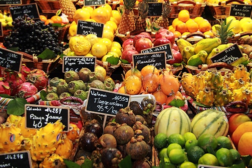 An assortment of colorful and exotic fruits displayed in baskets at a market. Each type of fruit is labeled with a small chalkboard indicating the name and price. Various fruits including pineapples, limes, dragon fruits, mangosteens, and pomegranates are visible.