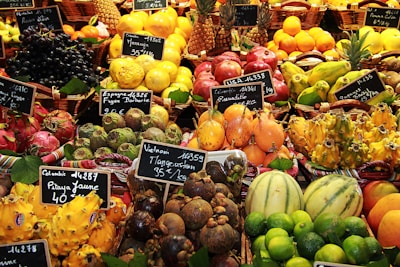 An assortment of colorful and exotic fruits displayed in baskets at a market. Each type of fruit is labeled with a small chalkboard indicating the name and price. Various fruits including pineapples, limes, dragon fruits, mangosteens, and pomegranates are visible.