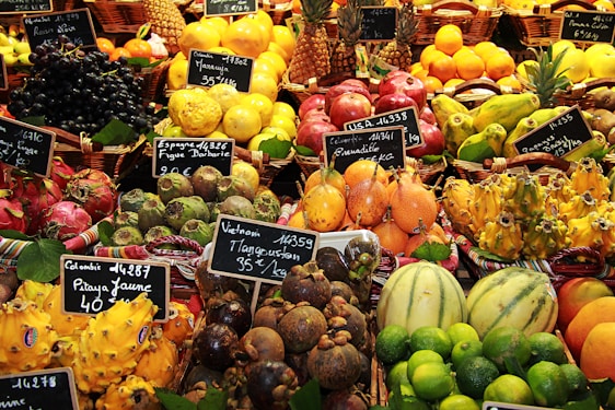 An assortment of colorful and exotic fruits displayed in baskets at a market. Each type of fruit is labeled with a small chalkboard indicating the name and price. Various fruits including pineapples, limes, dragon fruits, mangosteens, and pomegranates are visible.