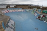 A skate park with a large, empty concrete bowl covered in colorful graffiti art along its edges. The scene is overcast with a cloudy sky and a body of water in the background, flanked by old brick buildings and stone walls.