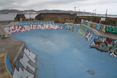 A skate park with a large, empty concrete bowl covered in colorful graffiti art along its edges. The scene is overcast with a cloudy sky and a body of water in the background, flanked by old brick buildings and stone walls.