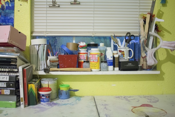 A cluttered workspace with various art and craft supplies arranged on a window sill. Items include containers of Mod Podge, paint, brushes, scissors, tape rolls, and a mug. The background has a window with closed blinds, and there's a yellow wall. Books and folders are stacked on the left side, and several paint cans and jars are on the table.